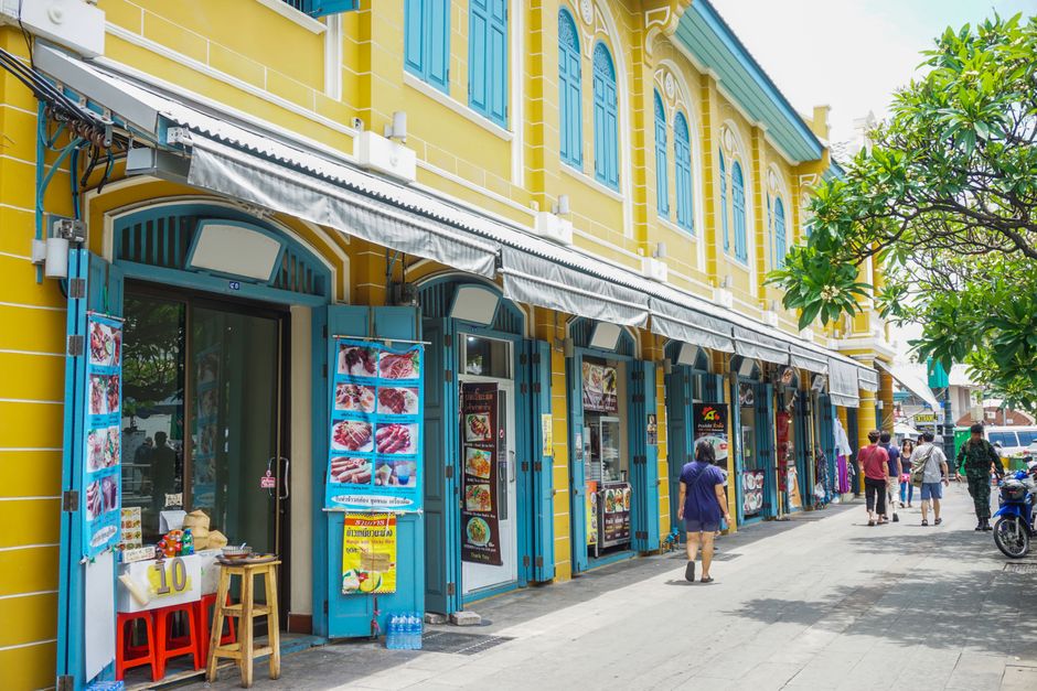 Vibrant street scene in Bangkok with people strolling past historic yellow and blue architecture.