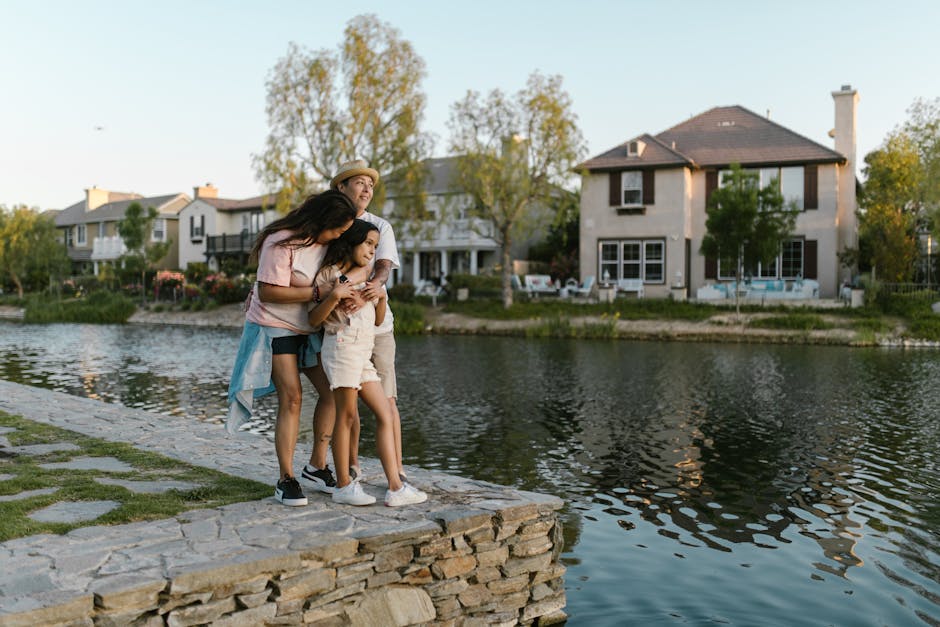 A joyful family embraces by a tranquil lake with beautiful houses in the background.