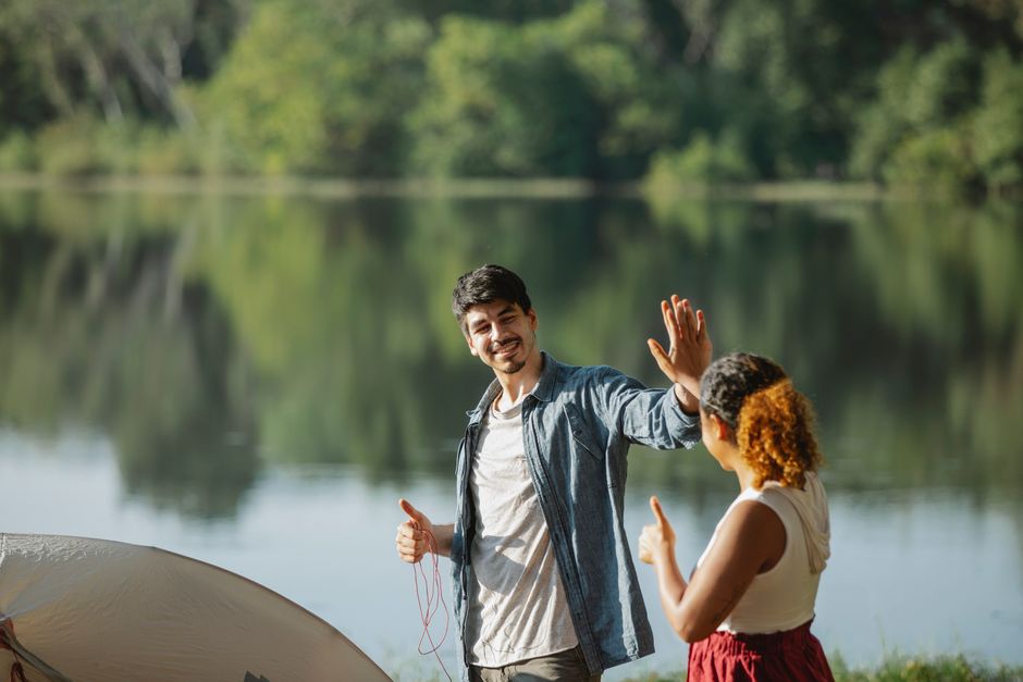 Happy young traveling couple giving each other high five while standing on river bank and putting up tent