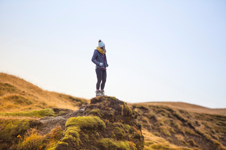 Woman in warm clothing standing on a moss-covered hilltop under a clear blue sky, embracing adventure and nature.