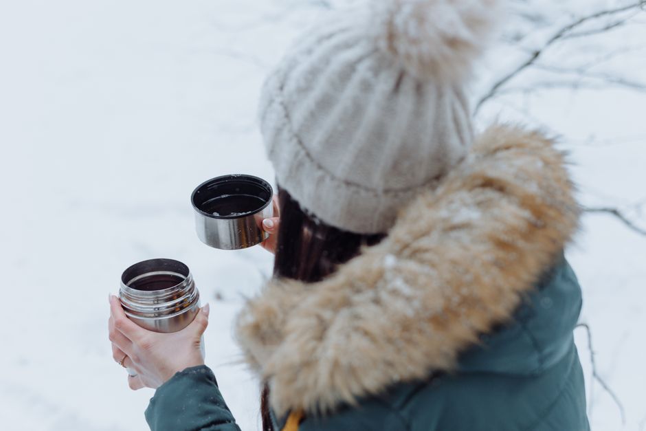 Woman holding thermos cup in snowy landscape, wearing warm winter clothing.