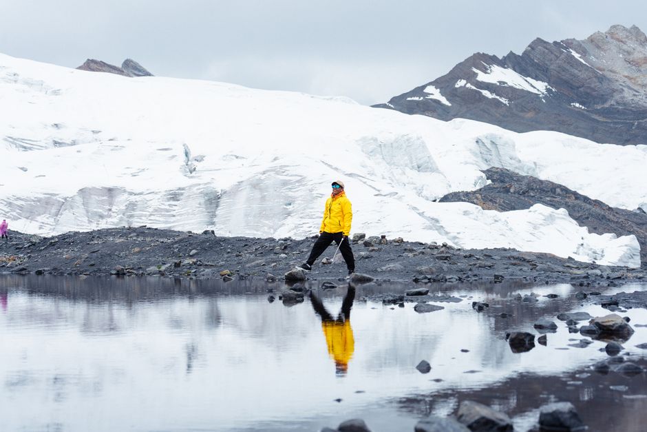 Person in yellow jacket walks by a reflective glacier pond, showcasing a serene and cold environment.