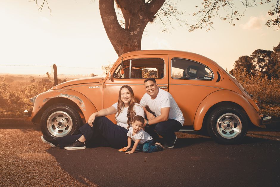 Happy family sitting by a classic orange car under a tree during sunset.
