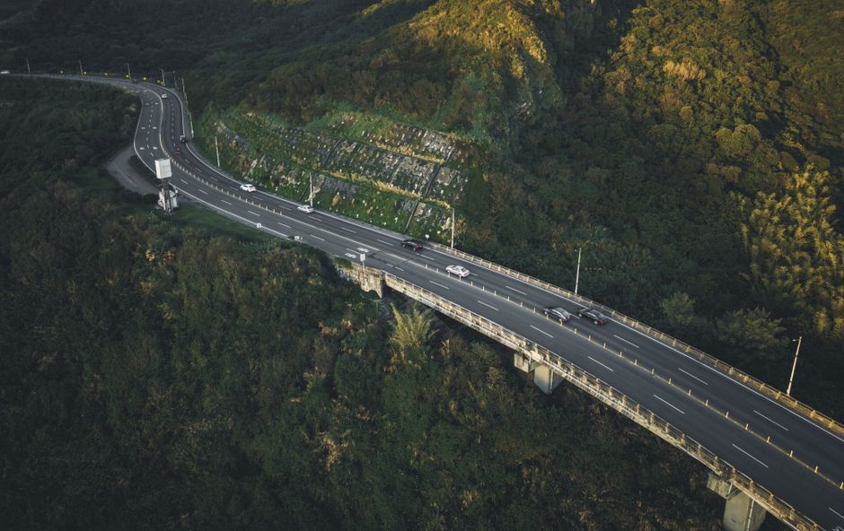 Aerial shot of a highway bridge cutting through dense, green hills in daylight.
