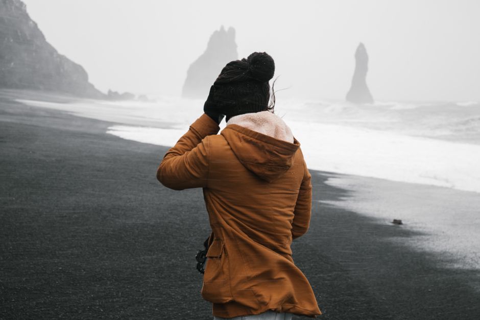 A woman walks on a windy beach with dramatic sea stacks in the distance, showcasing an adventurous coastal landscape.