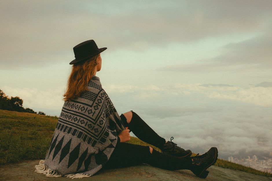 A young woman in a patterned poncho and hat sits on a hill, enjoying a panoramic view in Jaraguá do Sul, Brazil.