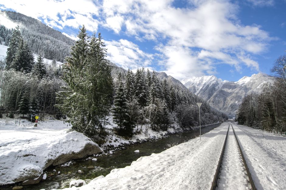 Scenic winter landscape featuring snow-covered alpine mountains, pine trees, a flowing river, and train tracks under a blue sky.
